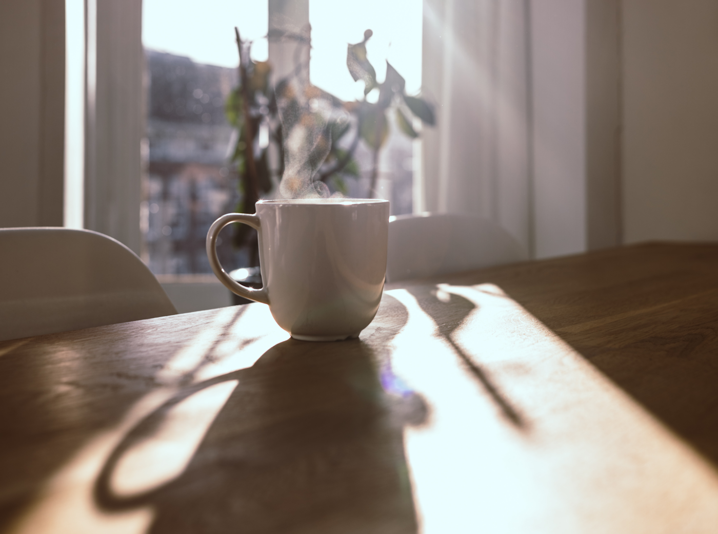 朝の光が差し込む窓辺の木のテーブルに置かれた、湯気の立つ白いマグカップのコーヒー